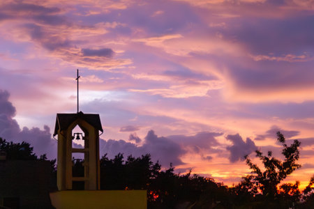 A stunning sunset fills the sky with hues of orange, pink, and purple. The silhouette of a church bell tower stands out against the colorful backdrop, surrounded by trees.の写真素材