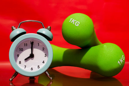 Small Alarm clock on the background of dumbbells for sports.