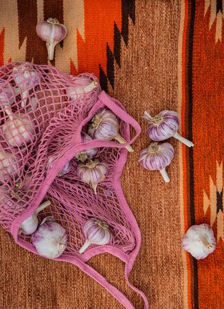 Above view of many pink and purple garlic in eco knitted bag on orange background. Big and little organic garlic on ethnic carpet on ground. Concept of food and harvesting.の写真素材