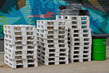 Stacks of white wooden pallets and green metal barrel in the background of painted brick wall