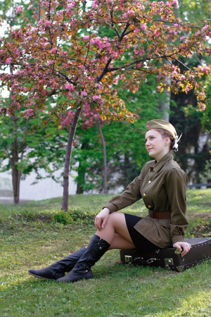 Pretty Soviet female soldier in uniform of World War II sits on a suitcase near flowering treeの写真素材