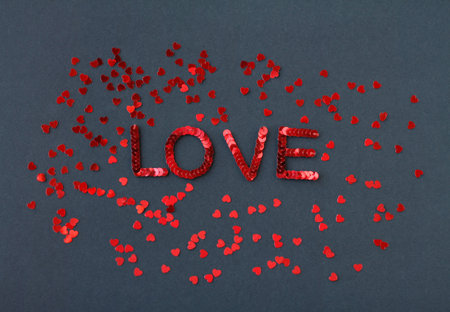 Inscription of "Love" embroidered with red sequins on black textured background with scattered random sequins in form of heartsの写真素材