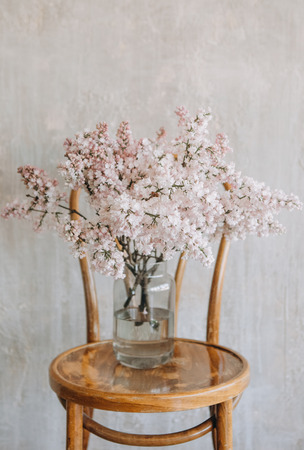 lilac flowers in a glass vase on a wooden chair against a textured wallの写真素材