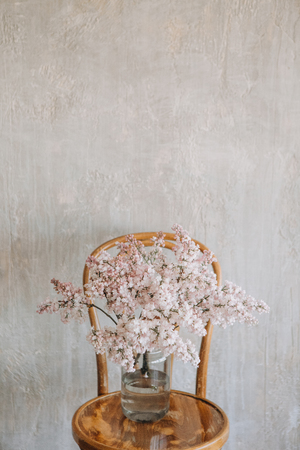 lilac flowers in a glass vase on a wooden chair against a textured wallの写真素材