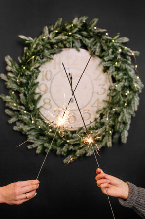festive sparklers in the hands of two women on the background of hours in a Christmas wreath