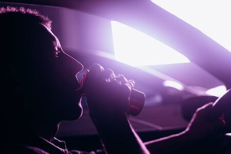 man drinks coffee from a jar in a car at night city and bright lightsの写真素材