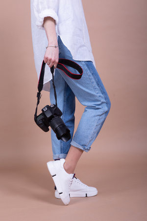 woman photographer in jeans and white sneakers holding a camera on a beige backgroundの写真素材