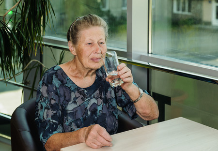 Elderly woman drinking water from a glass sitting at tableの写真素材