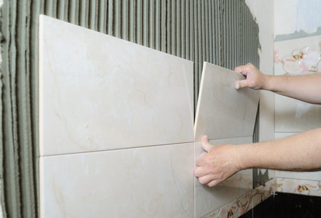 Tilers hands are  installing a ceramic tile on a wall in a bathroom.