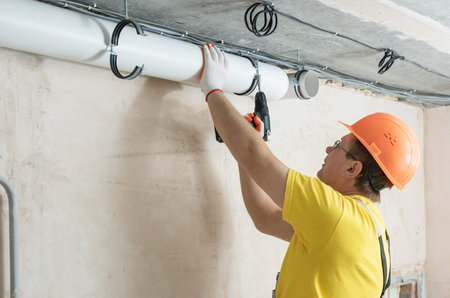 The worker is fixing ventilation pipes to the ceiling with a screwdriver.