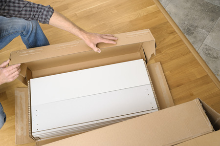 A man is unpacking a cardboard box with furniture. On the floor there is a box with white boards.の写真素材