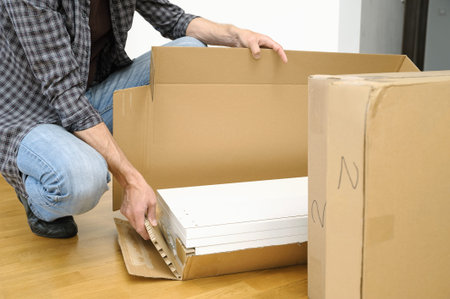 A man is unpacking a cardboard box with furniture. On the floor there is a box with white boards.