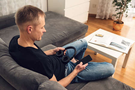 The man in headphones and with a smartphone sits on the couch. Back view.の写真素材