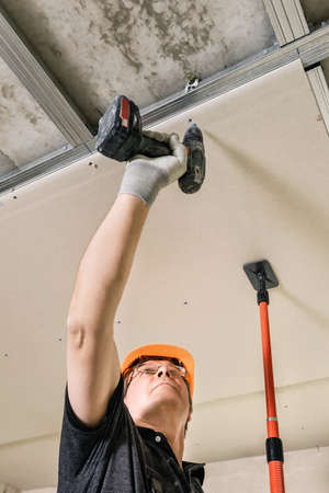Installation of drywall. Worker is using screws and a screwdriver to attach plasterboard to the ceiling.の写真素材