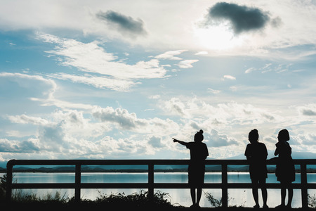silhouette of young girls stand at fence at sunset with light and lake view backgroundの写真素材