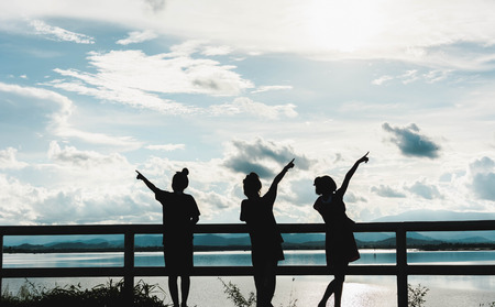 silhouette of young girls stand at fence at sunset with light and lake view backgroundの写真素材