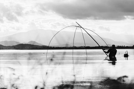 silhouette of fisher man use square dip net fishing at lake with mountain and blue sky background in white toneの写真素材