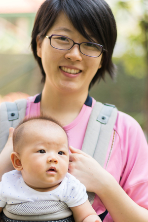 Asian  mother and her baby girl in a baby carrier, outdoors portraitの写真素材