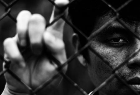Depressed man standing behind a fence, hand grabs steel mesh cage,close up on face in white tone, abuse conceptの写真素材
