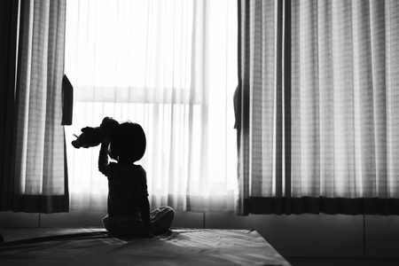 Little baby child sitting on bed playing with the doll in the dark room with light throught curtainの写真素材