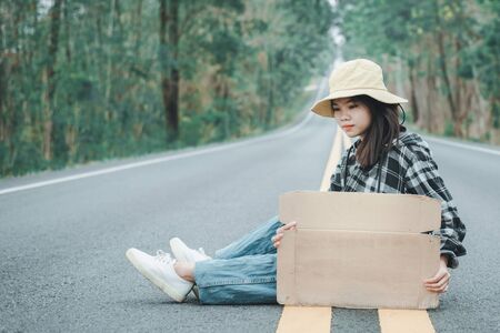 Travel hitchhiker woman holding sign with hat and backpack sitting on a road の写真素材