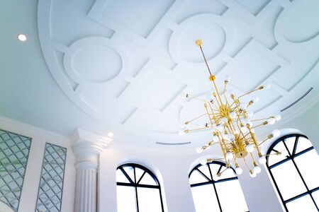 Chandelier with bulbs and brass hanging on the ceiling in a white room with windows.の写真素材