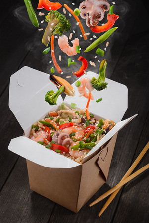 Rice with seafood and vegetables. Octopuses, mussels, shrimps, broccoli, pepper, lettuce, asparagus and sesame seeds are falling down into take-out box, eat using chopsticks.の写真素材