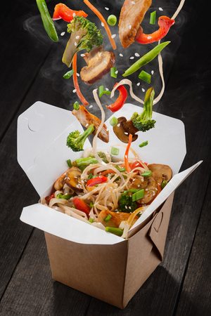 Chinese noodles with vegetables. Ingredients are falling into box: noodles, chicken meat, mushrooms, broccoli, pepper, lettuce, asparagus and sesame seeds. Presentative photo of food product.の写真素材