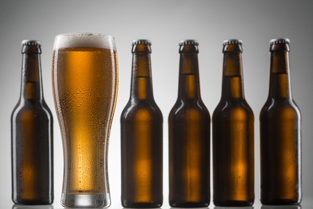Line of bottles and glass of chilled beer on neutral background. Focus on tall glass of beer with thick head.の写真素材