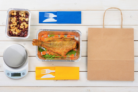 Paper bag, fish with arugula and sliced carrot, nuts and dried fruit, a food scale, plastic forks and knives on the wooden surface, a top-view image. Healthy takeout for two.の写真素材