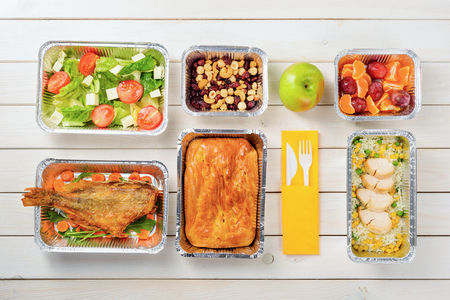 Fish, a pie, rice with corn and lean chicken, plums and tangerines, an apple, tomato and feta salad, hazelnuts, dried fruit and plastic cutlery on the wooden surface. Nutritional meal delivery.の写真素材
