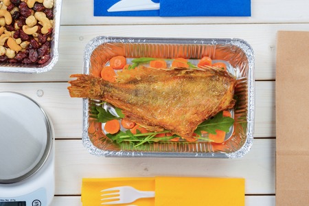 Appetizing fish with vegetables, cashews, hazelnuts and dried fruit, a kitchen scale and plastic flatware on the light wooden surface, close-up. Healthy takeout food.の写真素材
