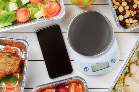 Kitchen scale, a smartphone on the wooden surface, close-up. Tomato and lettuce salad, cashews, hazelnuts and dried fruit, lean chicken and fish. Healthy delivery.の写真素材