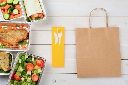 Plastic cutlery and a paper bag on the wooden surface. Tomato and feta salad, cheese sandwiches, fish with arugula, a vegetable roll, a sliced cucumber and baby carrots. Dinner food delivery.の写真素材