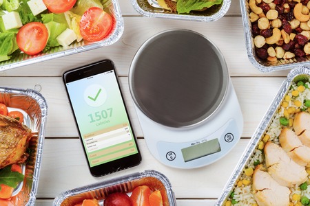 Calorie counter on the smartphone and food scale on the wooden surface. Lean chicken, nuts and dried fruit, tomato, feta and lettuce salad. Diet food delivery.の写真素材