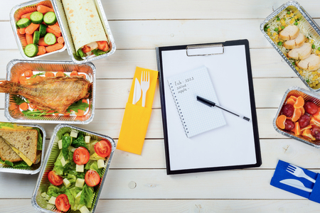 Note paper and a pen on the wooden surface. Plastic fork and a knife, fish with arugula, baby carrots, sandwich, tomato and feta salad, chicken, tangerines and plums. Counting calories.の写真素材