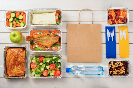Plastic cutlery, a paper bag, a bottle of water, fish, feta and lettuce salad, a pie, an apple, a vegetable roll, baby carrots, plums, tangerines, nuts and dates on the wooden surface. Order food.の写真素材