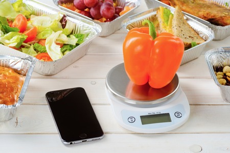 Kitchen scale and a fresh pepper, a smartphone on the light wooden table. Grapes, fish, cheese sandwiches, lettuce and tomato salad and a pie. Weight loss journey.の写真素材