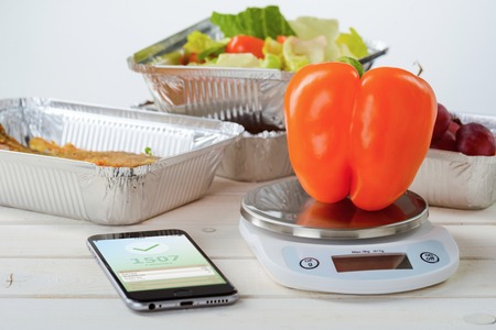 Fresh pepper on the food scale, calorie counter app on the smartphone, close-up. Lettuce salad and fish on the background. Healthy weight loss.の写真素材