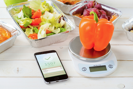 Calorie counter app on the smartphone, a kitchen scale and a fresh pepper on the wooden surface, close-up. Lettuce and tomato salad, grapes on the background. Counting calories.の写真素材