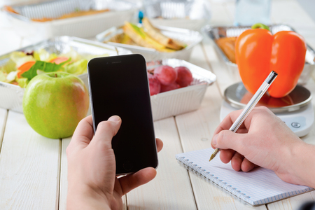 Holding a smartphone and making notes, close-up. Kitchen scale, a pepper, an apple, grapes and various dishes on the background, a light wooden table. Food diary.の写真素材
