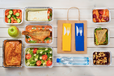 Bottle of water, forks and knives, a paper bag, fish, tomato and feta salad, a pie, a vegetable roll, baby carrots, an apple, fruits, a sandwich, nuts and dates on the wooden surface. Takeout food.の写真素材