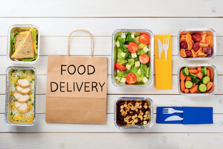 Paper bag with Food Delivery sign. Cashews, hazelnuts and dates, carrots and cucumbers, rice with chicken, sandwiches, tomato salad, plastic cutlery and fruit, wooden surface. Ordering your meal.