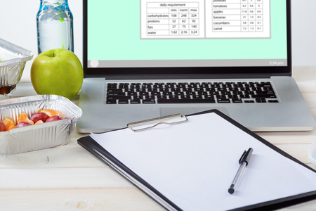 Laptop with nutrient calculations, note paper and a pen, a bottle of water, an apple, plums and tangerines, a white table, close-up. Dietitian career.の写真素材