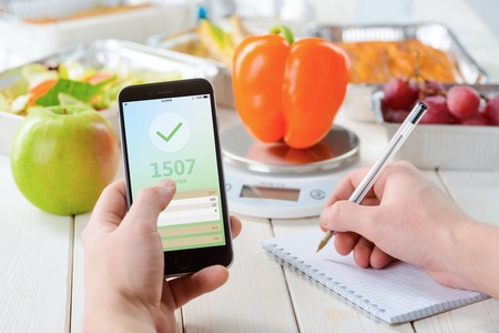 Calorie counter app on the smartphone, making notes, close-up. Grapes, an apple on the wooden surface, a pepper on the food scale on the background. Weight loss journey.の写真素材