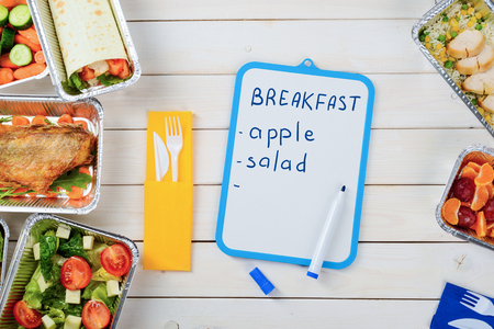 Dry erase board with food list and a marker on the wooden surface. Plastic cutlery, tomato and lettuce salad, fish, a vegetable roll, sliced cucumber, chicken, plums and tangerines. Meal plan.の写真素材