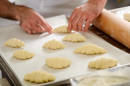 Man preparing croissants for baking, placing them on a tray. Bakery goods, snacks for coffee or tea.の写真素材