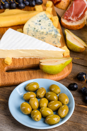 Italian cheeses, meat and fruits. Focus on a plate of green olives, grape, pear halves, cheese and toasts with jamon in blurred background. Cooking a delicious meal for lovers.の写真素材
