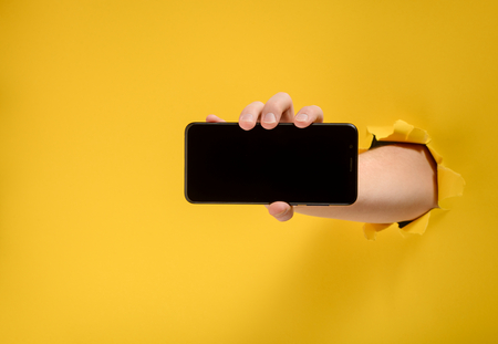 Hand holding a cell phone, showing a blank screen through torn yellow paper background. Gadget mockup.の写真素材