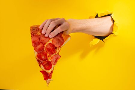Hand holding a slice of Pepperoni pizza through a torn hole in yellow paper background. Have a snack, fast food advertisement.の写真素材